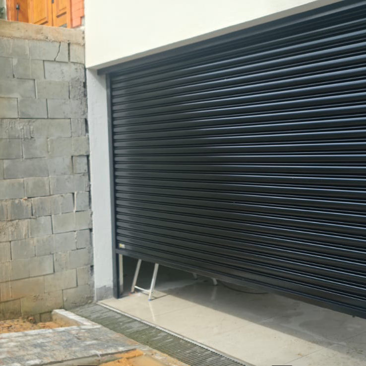 Portão de enrolar preto, largo, numa garagem com parede de bloco
