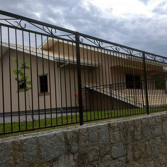Grade preta com arabescos sobre muro de pedra, com a casa ao fundo